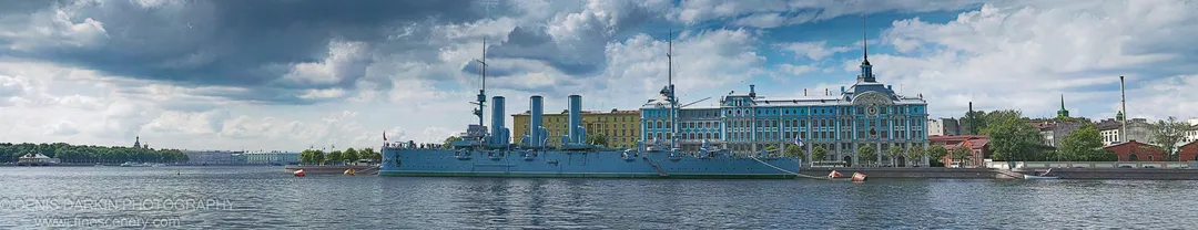 A historic battleship moored alongside an ornate blue building on the Neva embankment in St Petersburg, storm clouds gathering overhead.