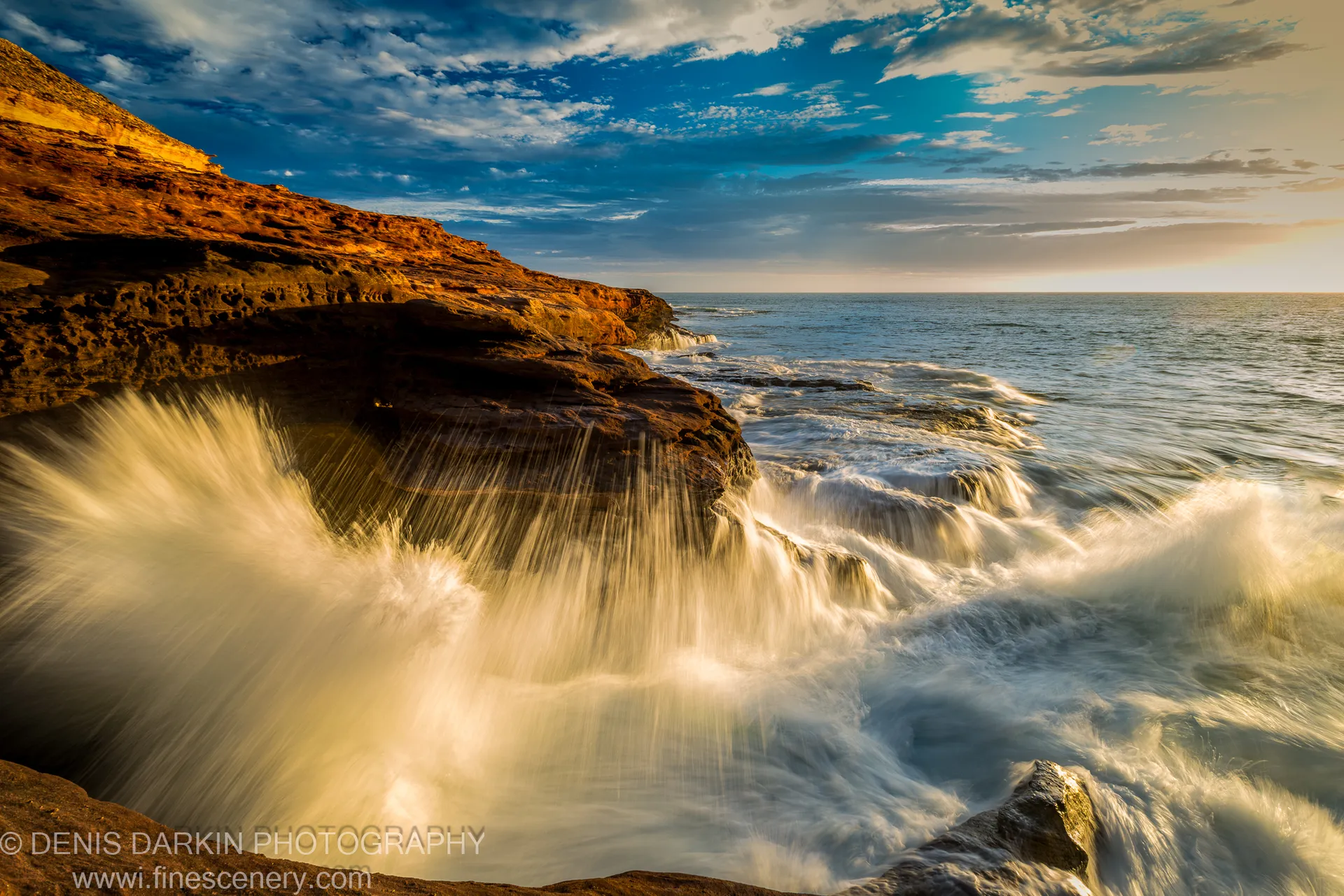 Waves crash against the iron-rich sandstone base of Red Bluff, the surf blurred to white foam by a long exposure.