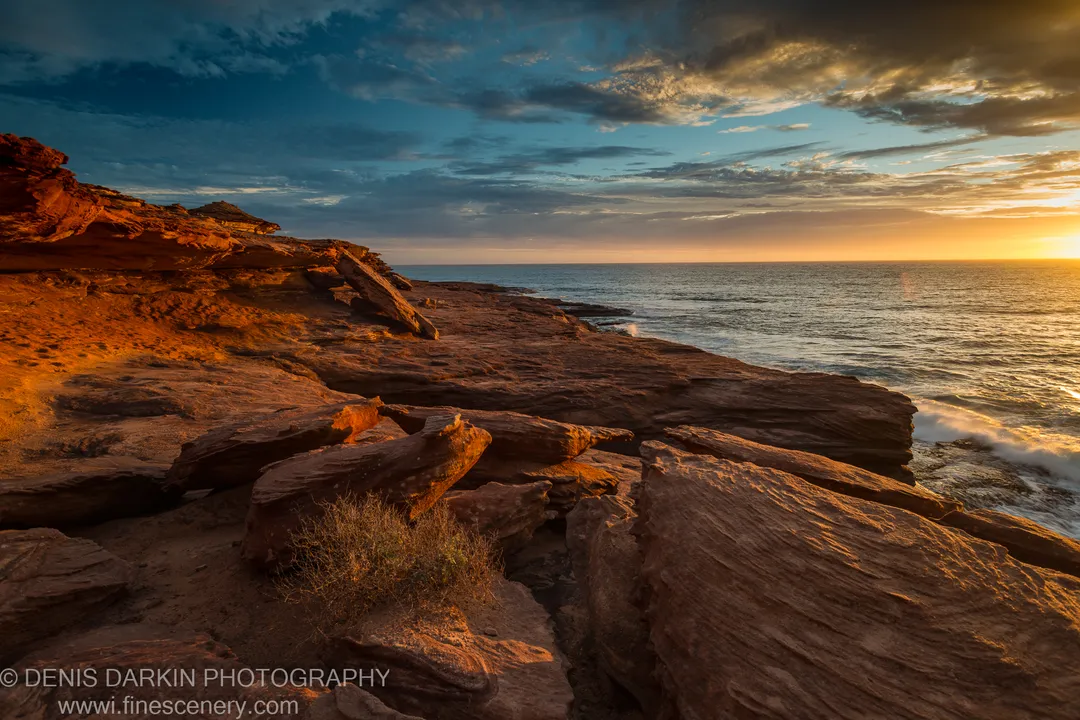 Eroded sandstone formations extend along the Kalbarri coast as evening light catches the warm red rock against a clearing sky.