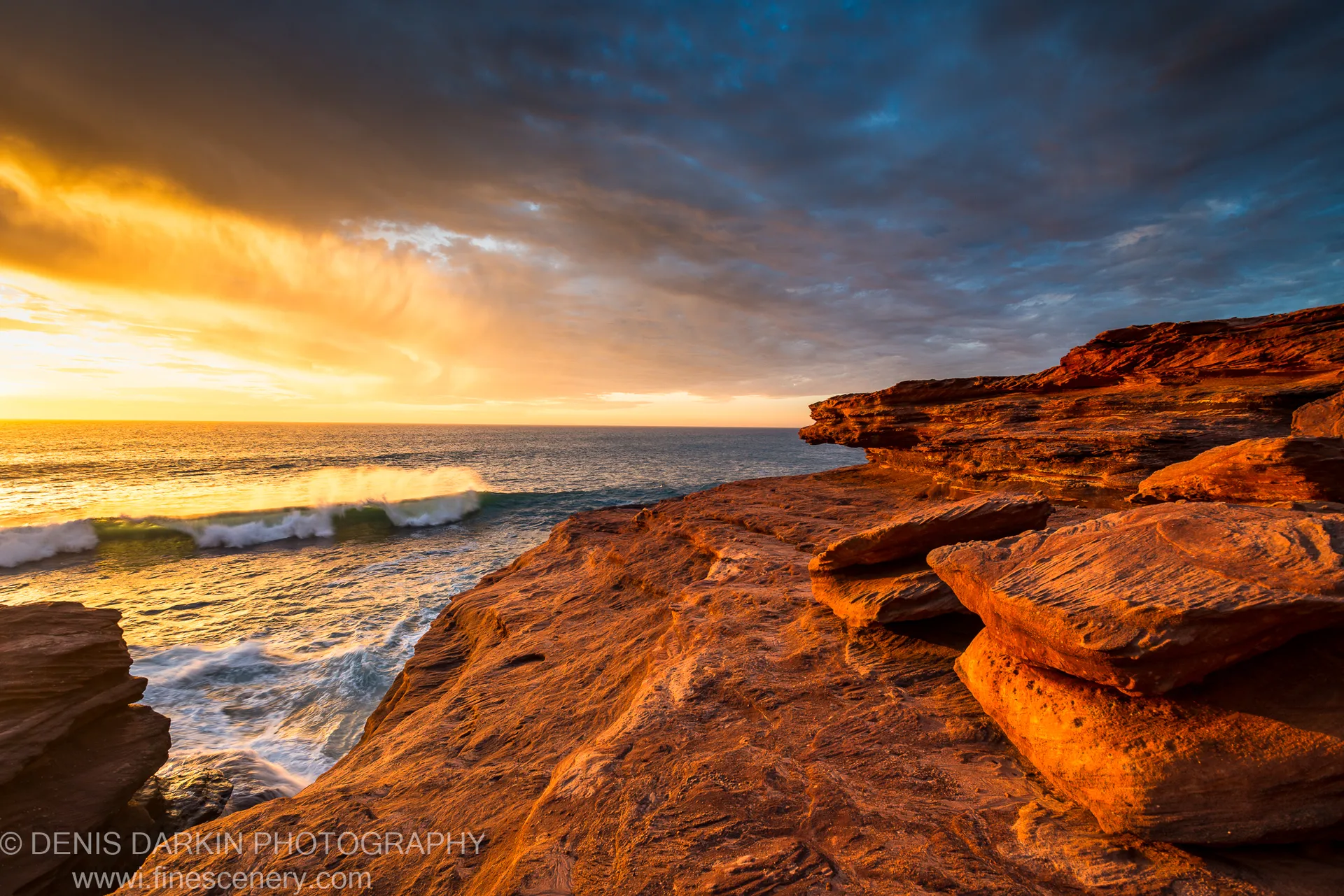 Late evening light warms the layered sandstone cliffs at Red Bluff as a storm retreats and white foam breaks at their base.
