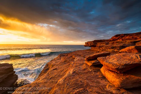 Late evening light warms the layered sandstone cliffs at Red Bluff as a storm retreats and white foam breaks at their base.