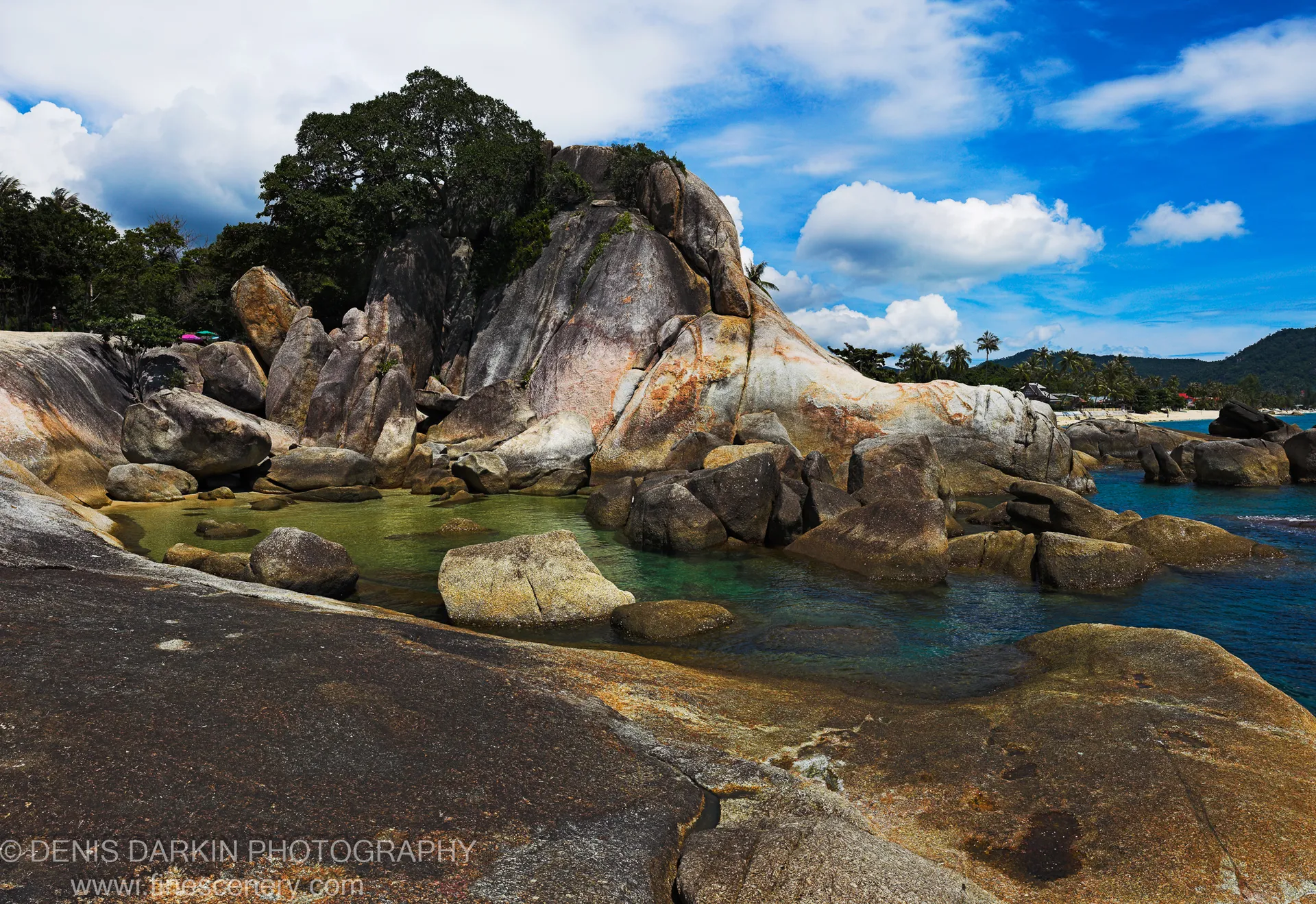 Smooth, rounded granite boulders surround a calm turquoise pool on the Thailand coast, with a broad canopy tree rising above against scattered clouds.