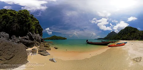 Traditional long-tail boats and colorful kayaks rest near a curving shoreline of turquoise water below lush karst hills on the Thai coast.