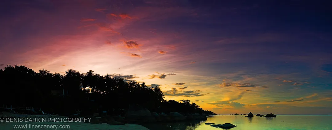 Silhouetted vegetation and rounded boulders line a calm shore at dusk in Thailand, with the evening sky reflected in still water below.