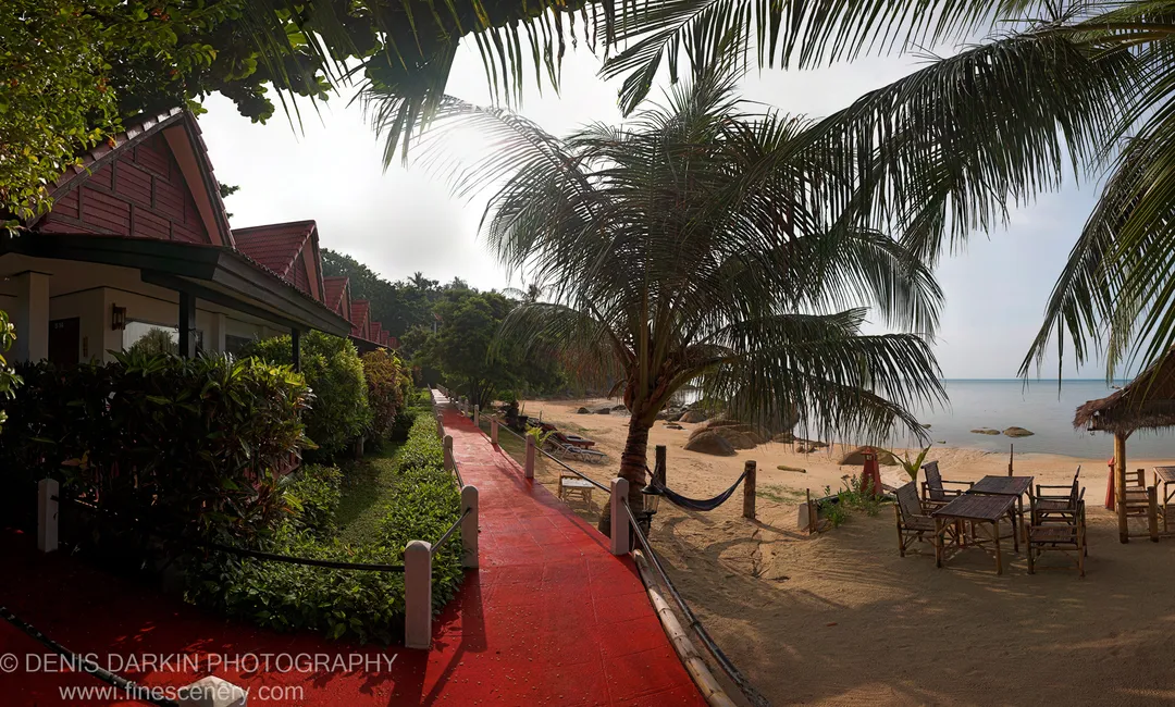 A terracotta-tiled walkway leads from a shaded tropical garden through overhanging palm fronds to a sunlit sandy beach and calm ocean beyond.