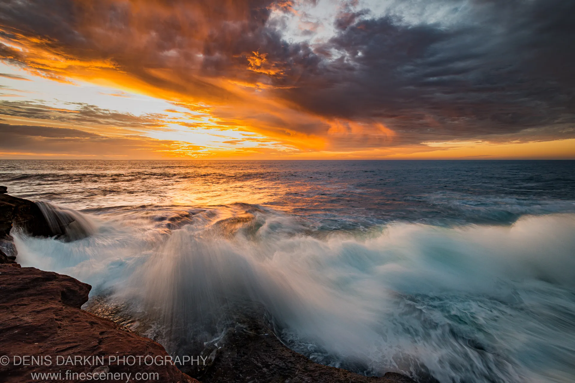 White water breaks over a red sandstone ledge at Kalbarri as an orange horizon lights the ocean surface beneath dark storm clouds.
