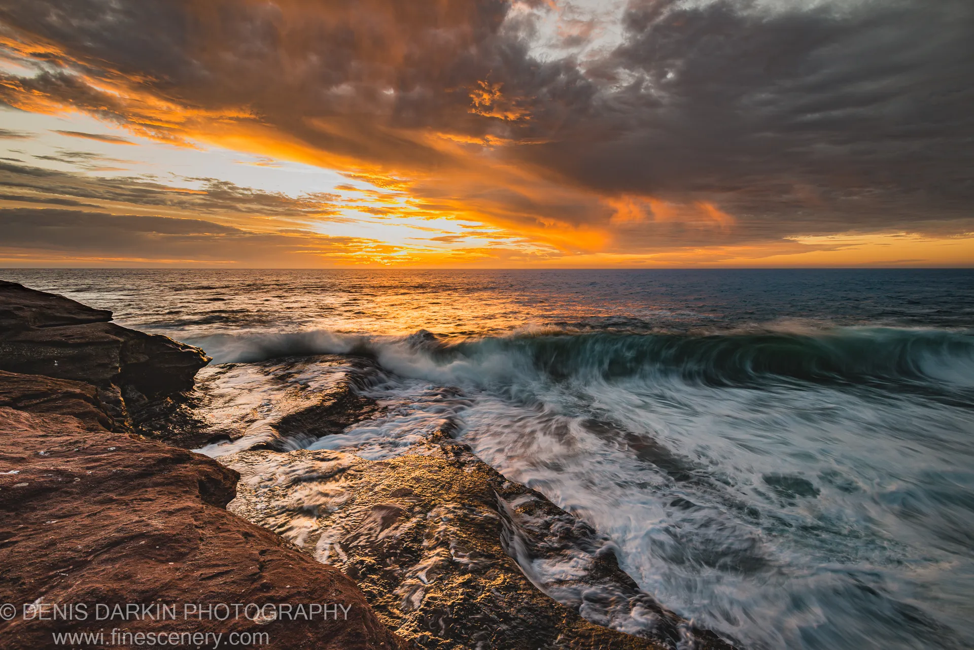 Long-exposure waves streaking over a wet rock platform on the Western Australian coast, with warm light filtering through heavy storm clouds in the evening sky.