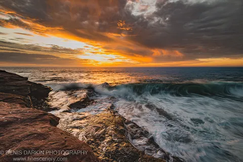 Long-exposure waves streaking over a wet rock platform on the Western Australian coast, with warm light filtering through heavy storm clouds in the evening sky.