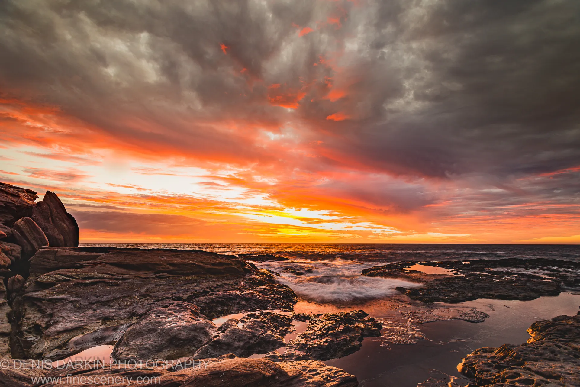 Layered dark rock formations and tide pools on the Western Australian coast, with the underside of storm clouds lit orange at evening.