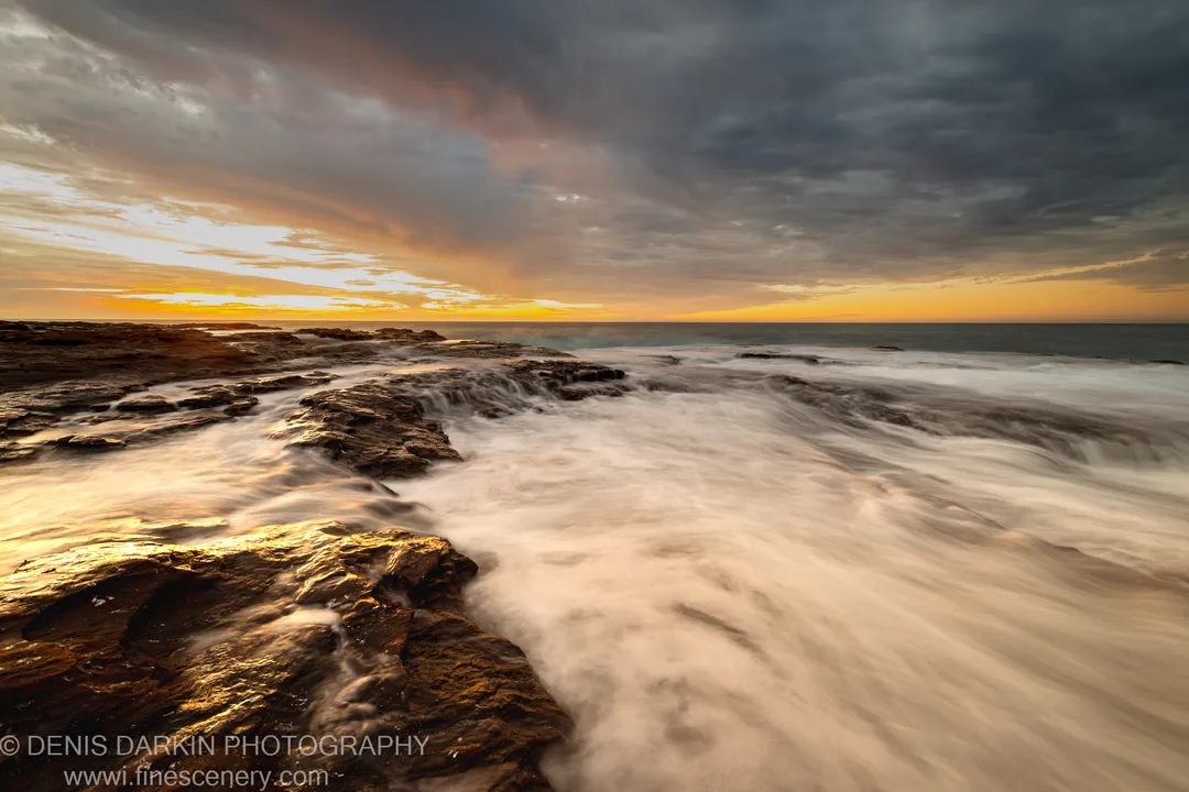 Long-exposure view of wave-washed basalt rocks on the Western Australia coast at sunset, with storm clouds above a golden horizon.