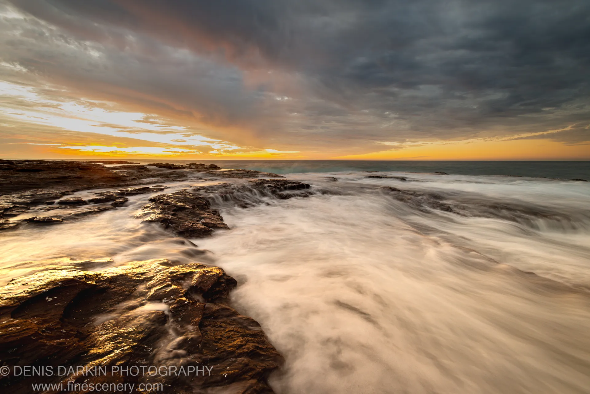 Long-exposure view of wave-washed basalt rocks on the Western Australia coast at sunset, with storm clouds above a golden horizon.