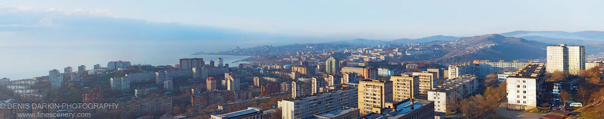 Dense mid-rise apartment blocks on hillsides in Vladivostok, lit by warm low-angle sunlight, with a coastal expanse and rolling hills in the distance.