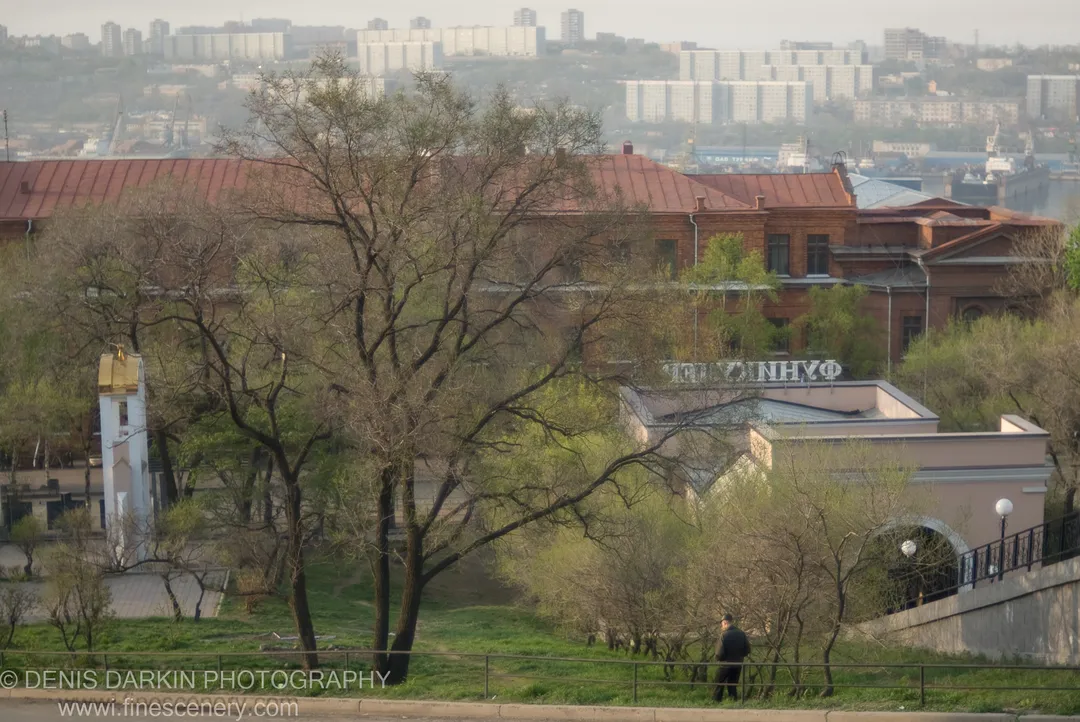A lone figure walks along a grassy park slope in early spring, with a red-brick building and hazy industrial waterfront visible behind in Vladivostok.