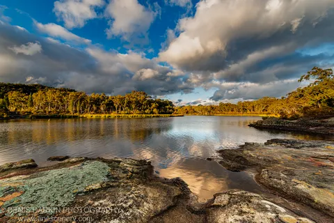 Lichen-covered rocks on the shore of Dunns Swamp, New South Wales, reflect a patchy cumulus sky over calm water in late afternoon.