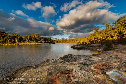 Reddish sandstone ledges extend along a river bend at Dunns Swamp, with sunlit eucalyptus trees on the far bank under scattered clouds.
