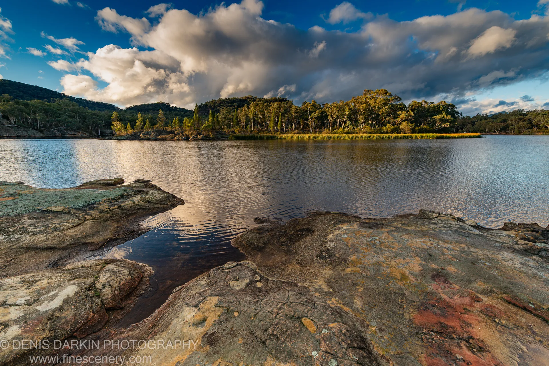 Weathered, iron-stained rock formations line the shore of a calm inland lake at Dunns Swamp, mirroring a wide sky of cumulus clouds.