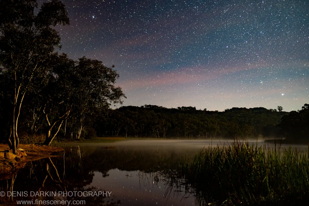 The Milky Way arches over the lake at Dunns Swamp, its faint light reflected in still water below silhouetted eucalyptus trees.